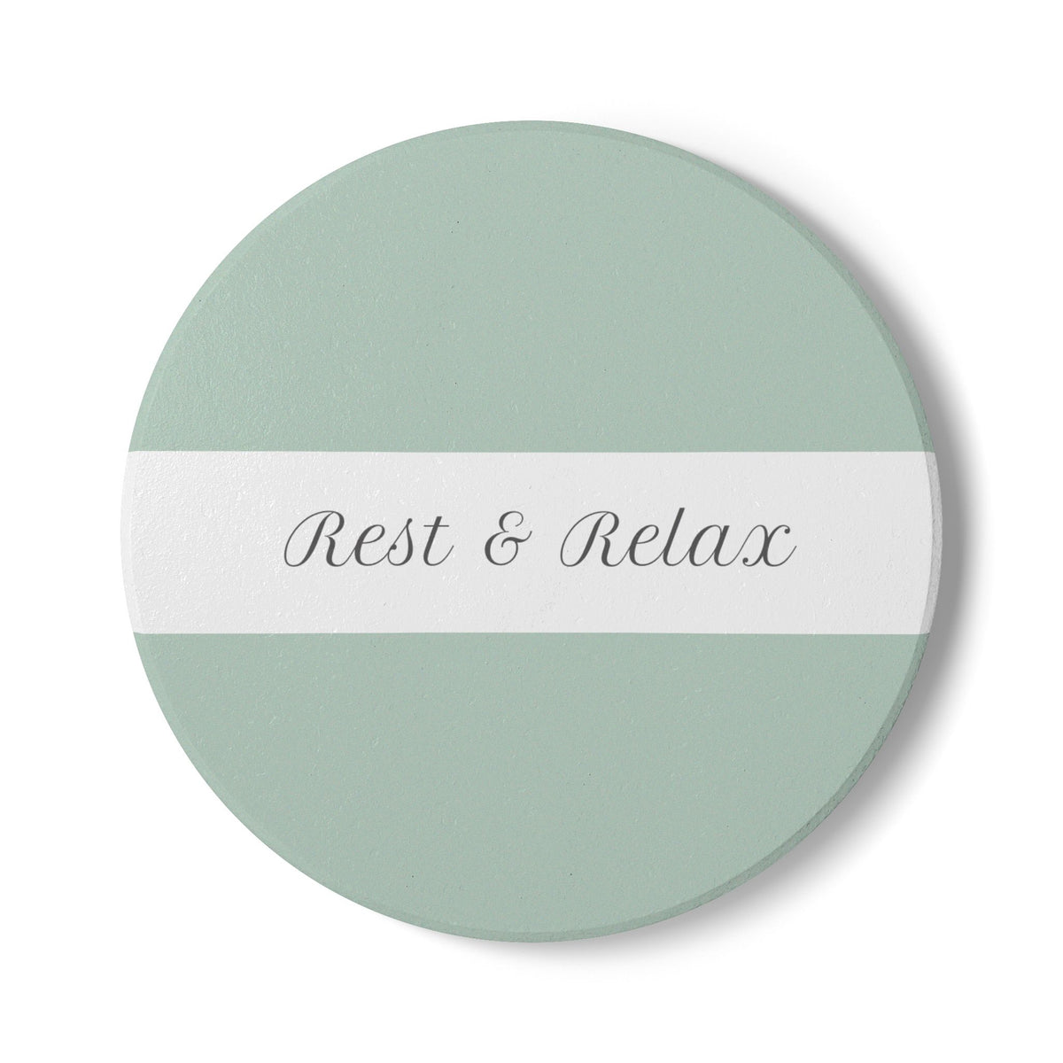 A round coaster with a sage green top and bottom half and a white horizontal center stripe. The text 'Rest & Relax' is printed in a gray cursive font across the stripe.