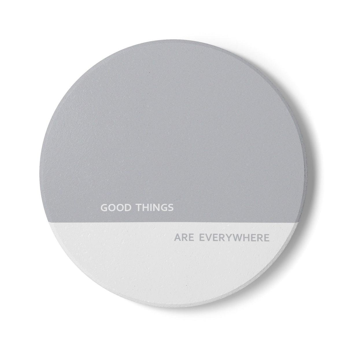 A round gray coaster with a white bottom half, featuring the printed text 'GOOD THINGS ARE EVERYWHERE' in a clean sans-serif font, shown on a white background with a soft drop shadow.