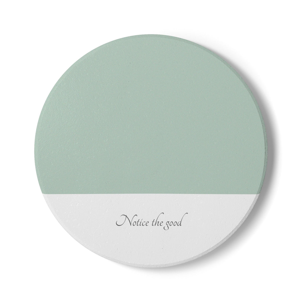 A round coaster with a split design, featuring a sage green top half and a white bottom half with the cursive text 'Notice the good'.