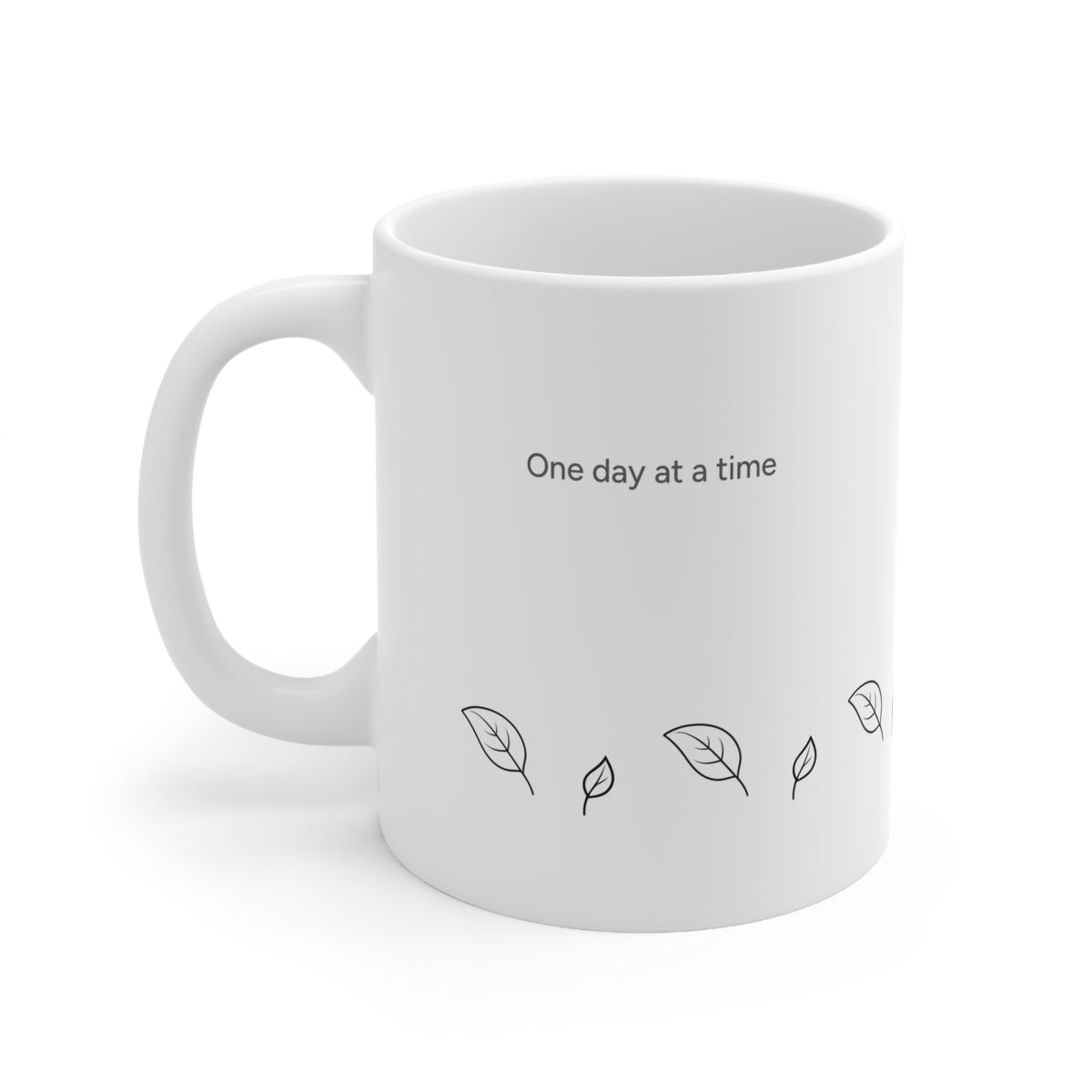Left side view of 11oz white ceramic coffee mug with the printed text 'One day at a time' and small black leaf illustrations, shown on a clean white background.