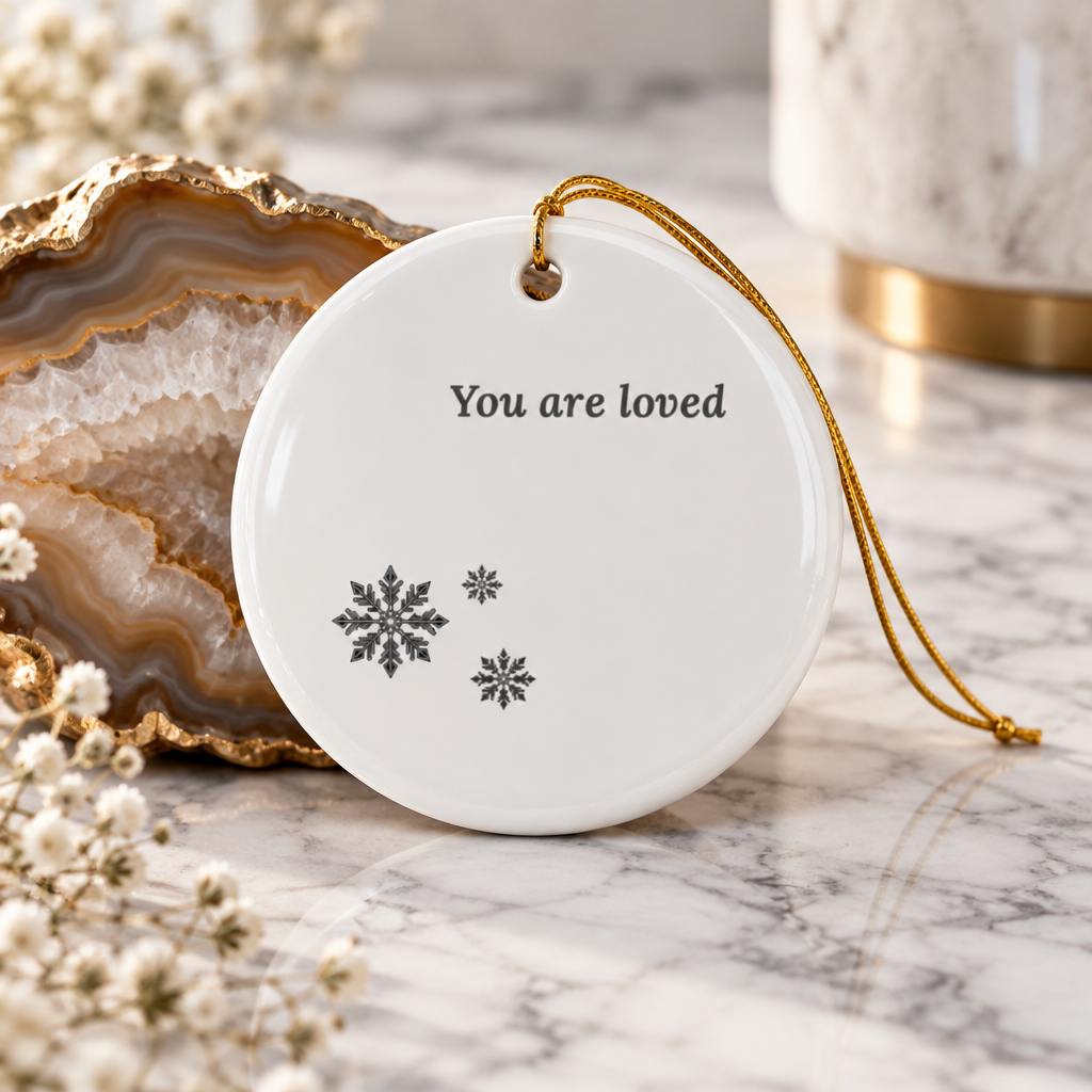 A round white ceramic Christmas ornament with the words 'You are loved' and three snowflake designs, hanging by a gold string on a marble surface with decorative props.