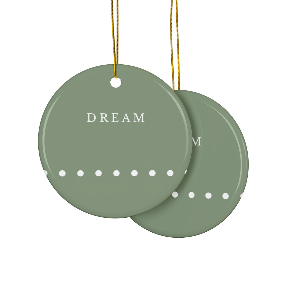 Two round sage green hanging ornaments with gold strings, one in front of the other. The front ornament is printed with the word 'DREAM' in white serif letters and a row of small white dots along the bottom.