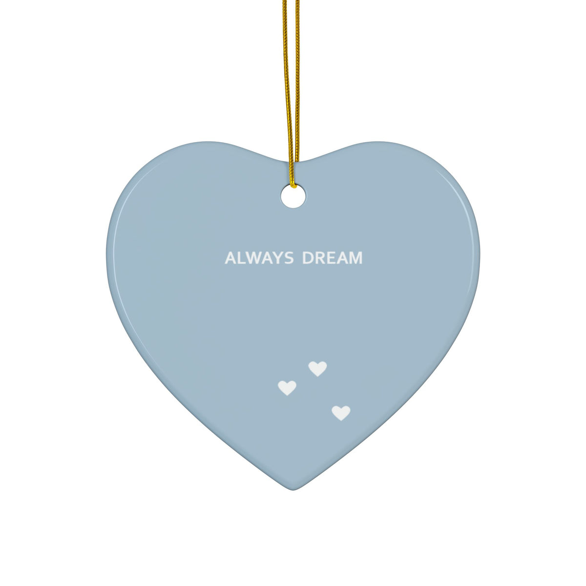 A heart-shaped ceramic ornament in a dusty light blue color, featuring the text 'ALWAYS DREAM' in white and three small white hearts near the bottom. It hangs from a gold string against a solid white background.