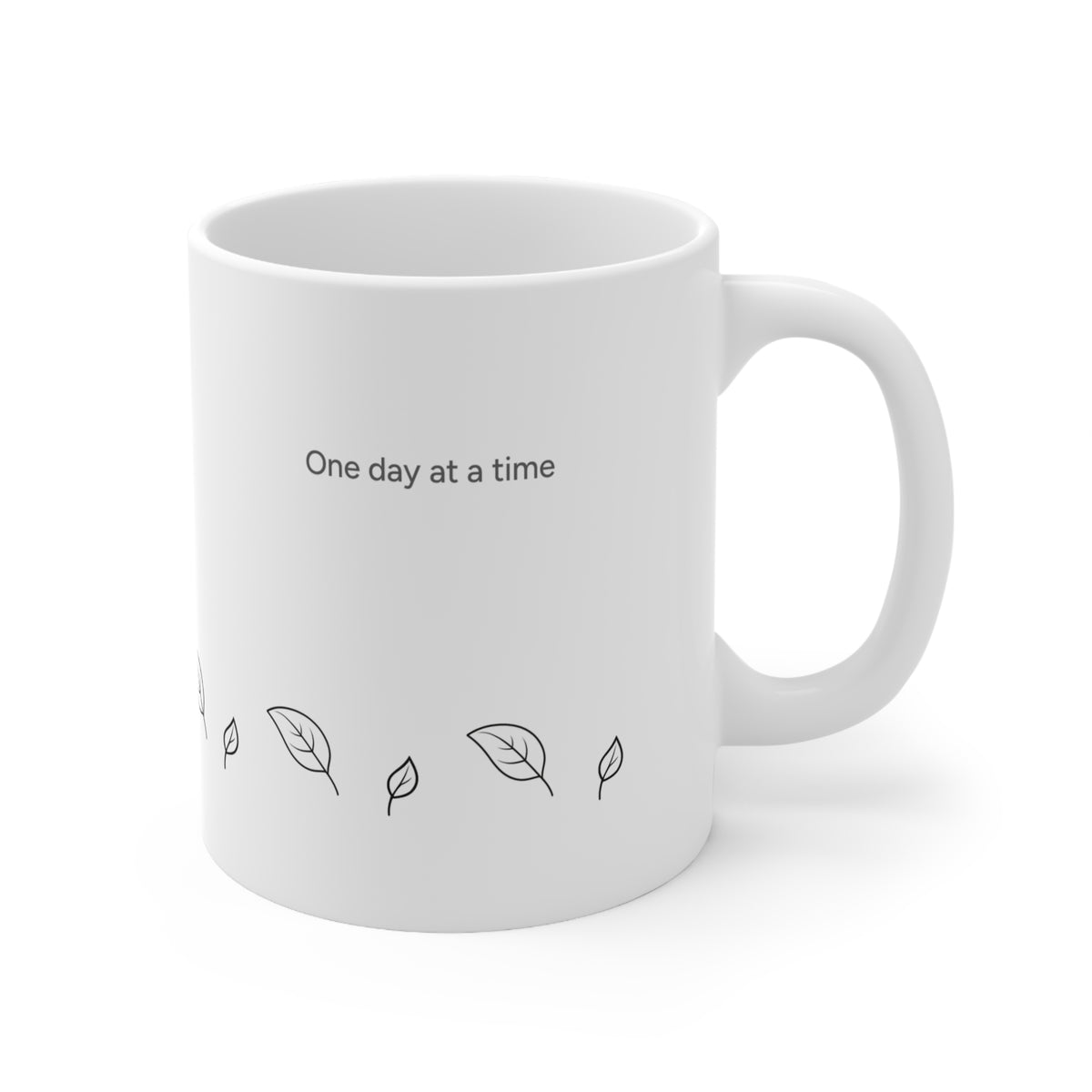 Right side view of 11oz white ceramic mug with the text 'One day at a time' printed in black near the top, and small black leaf line drawings along the lower front. The mug is shown on a clean white background with a soft shadow.