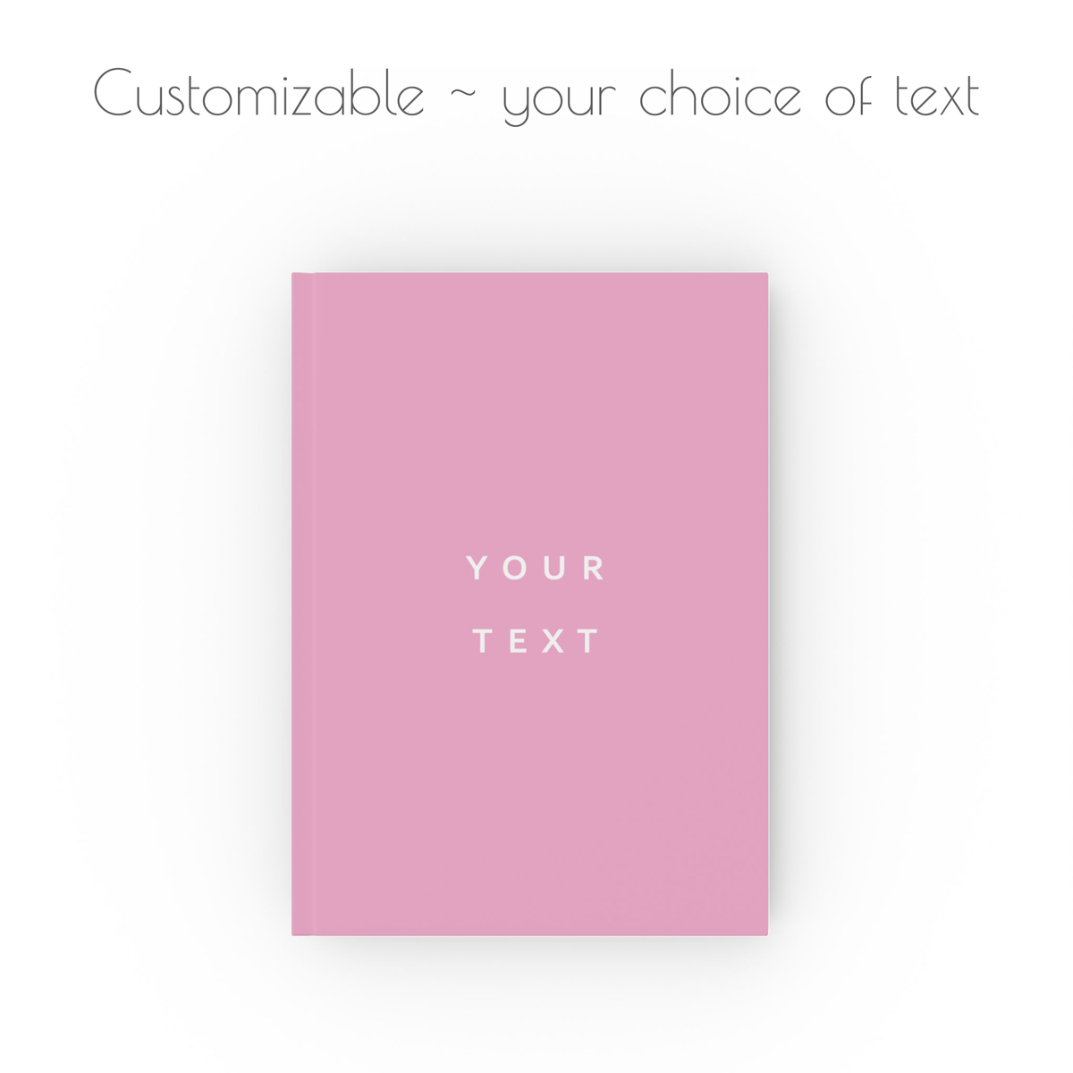 A pink hardcover notebook journal centered on a white background, with the words 'YOUR TEXT' printed in white in the middle. Above it, faint gray text reads 'Customizable ~ your choice of text'.