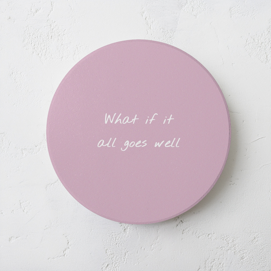 Pink round ceramic coaster with handwritten style text ‘What if it all goes well’ in white font. The coaster is on a grey cement style surface.