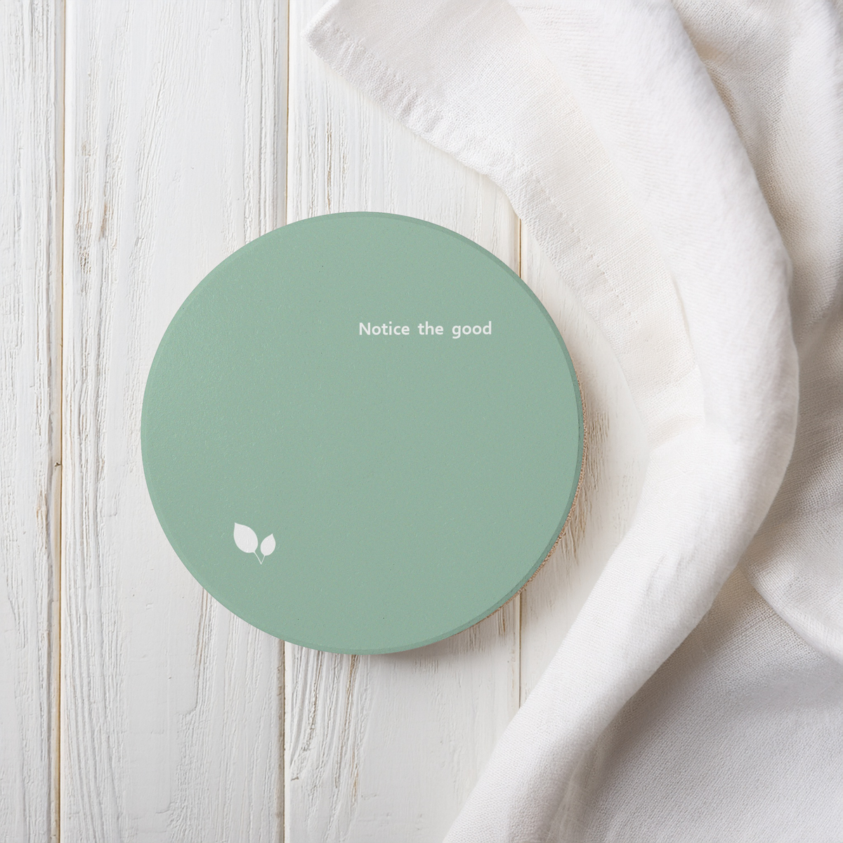 Green round ceramic coaster with delicate leaf motif design, and small, modern style text ‘Notice the good’ in white font. The coaster is on a white wooden surface next to linen.