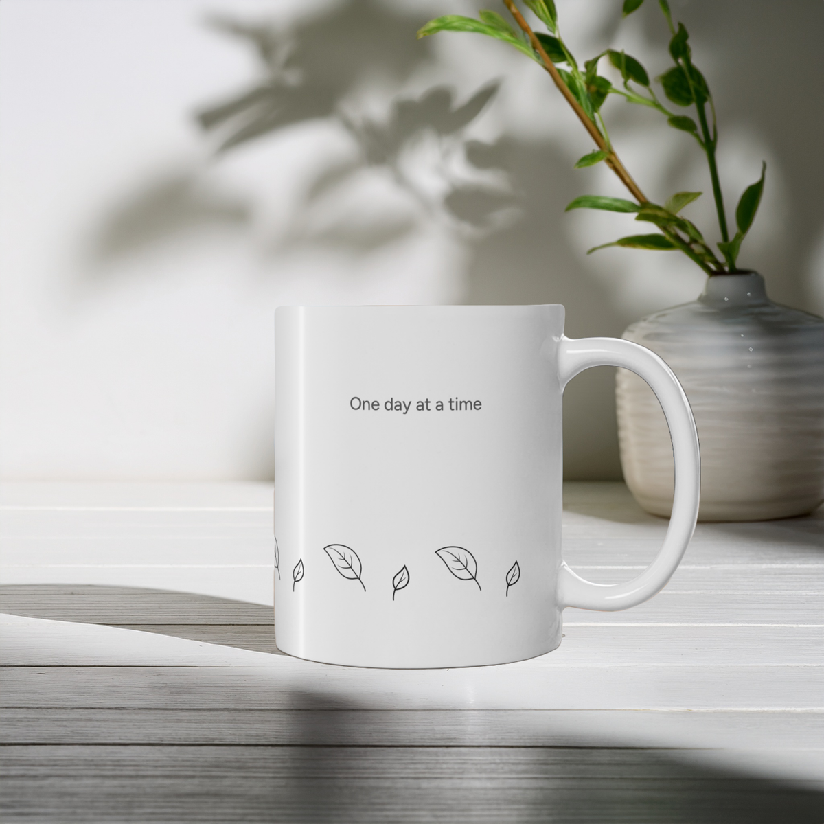 An 11oz white ceramic mug with the printed text 'One day at a time' and small leaf illustrations, placed on a white tabletop with soft shadows. A gray vase with green stems sits in the background.