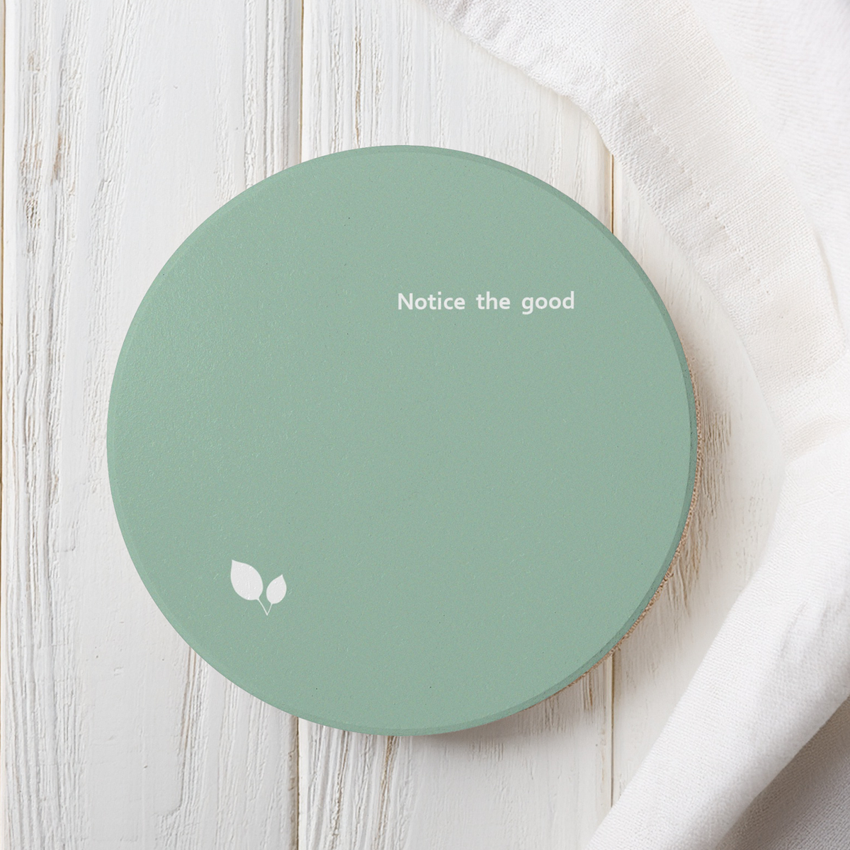 Green ceramic coaster with small white leaf motif and small white text 'Notice the good'. The coaster sits on a white wooden surface next to linen.