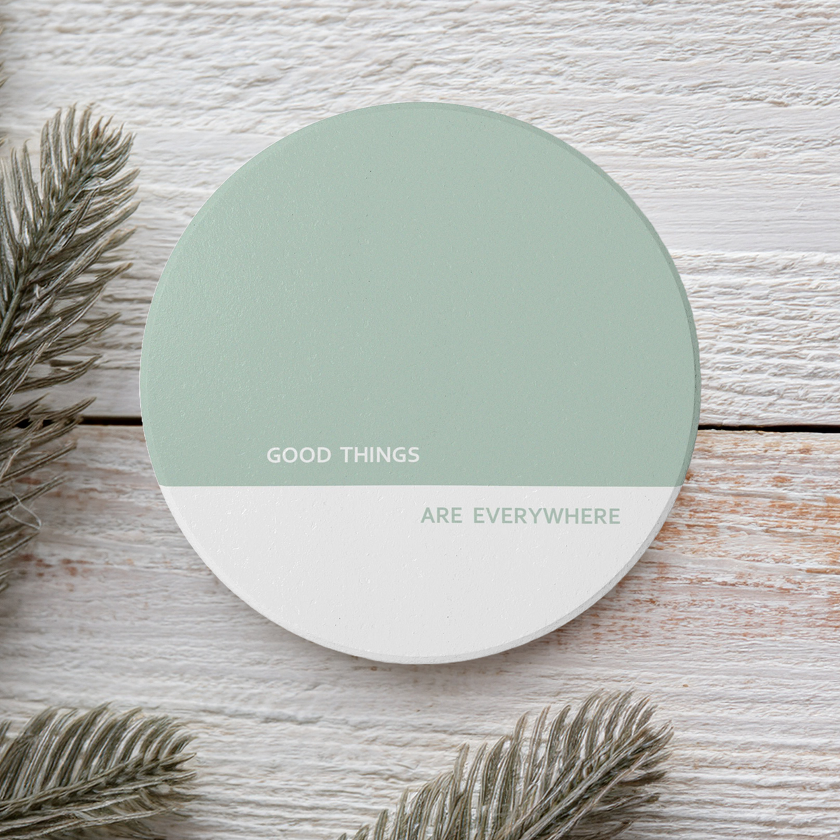 A round coaster with a split design, featuring a sage green top half and a white bottom half with the text 'GOOD THINGS ARE EVERYWHERE' in white. The coaster is styled on a white-washed wooden background with pine needles.