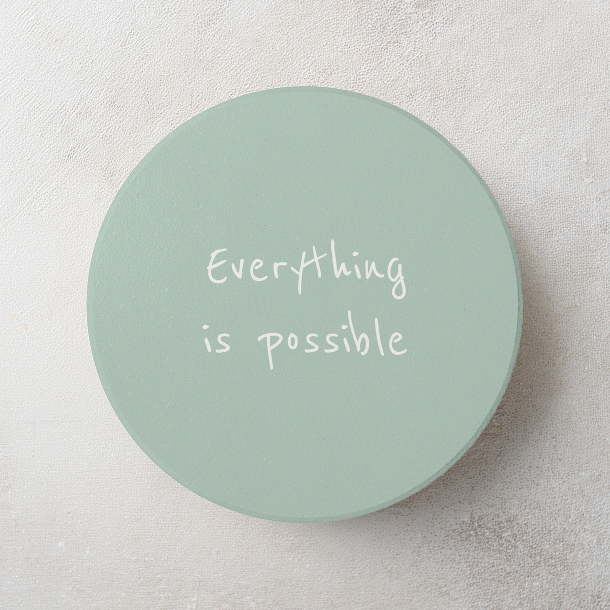 A round, light sage green coaster with the phrase 'Everything is possible' printed in white handwritten-style text, displayed on a textured light gray surface.