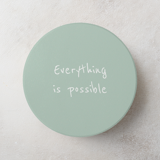 A round, light sage green coaster with the phrase 'Everything is possible' printed in white handwritten-style text, displayed on a textured light gray surface.