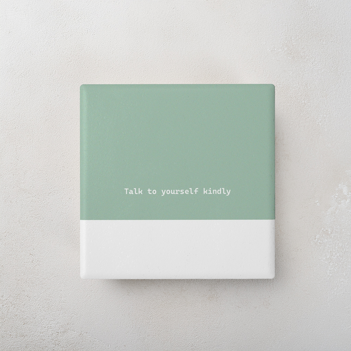 Green and white square ceramic coaster with modern style text ‘Talk to yourself kindly’ in white font. The coaster is on a cement style surface.