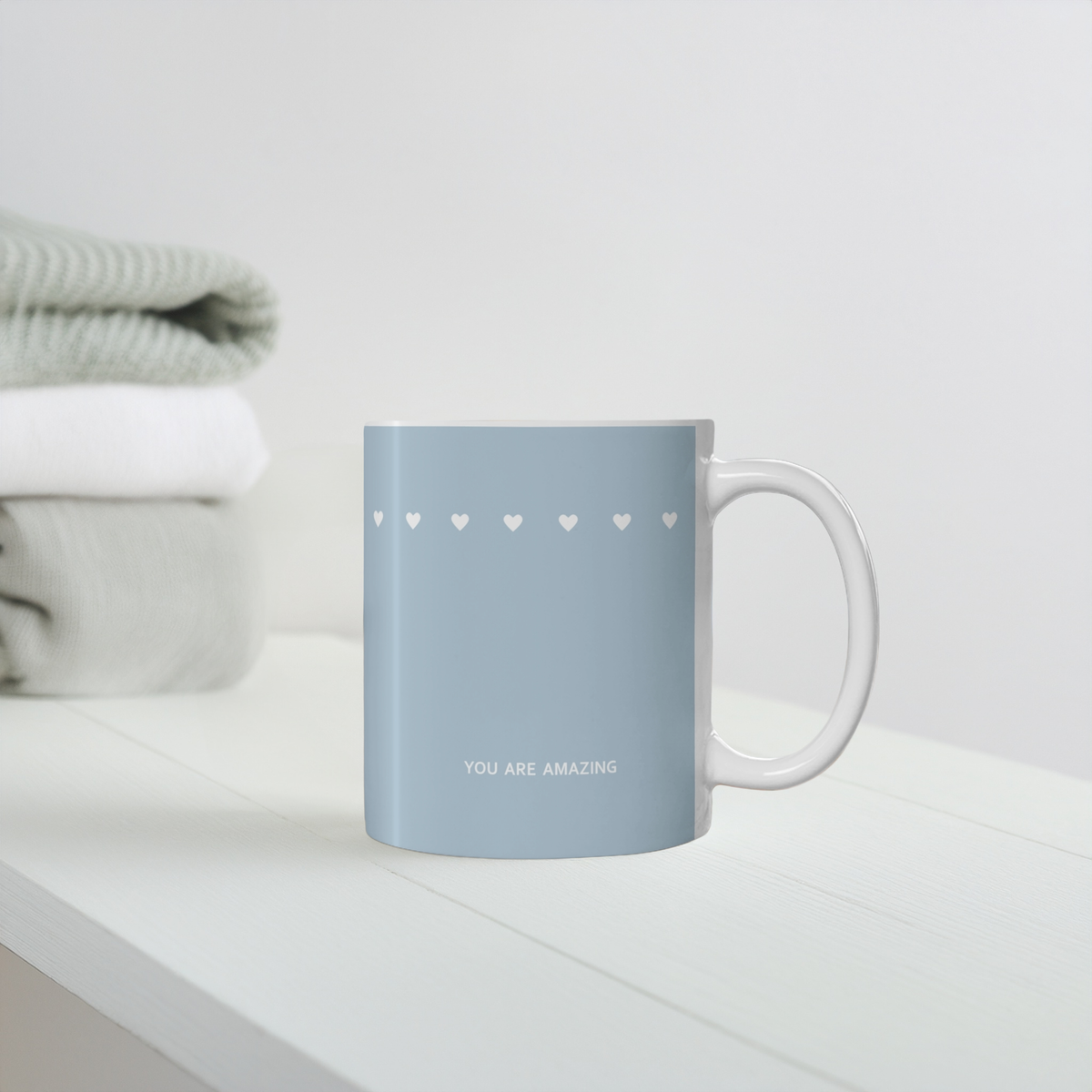 Blue ceramic mug with row of small white hearts. Small white font at the bottom ‘You are amazing’. The mug sits on a white wooden shelf with cozy folded jumpers in background.
