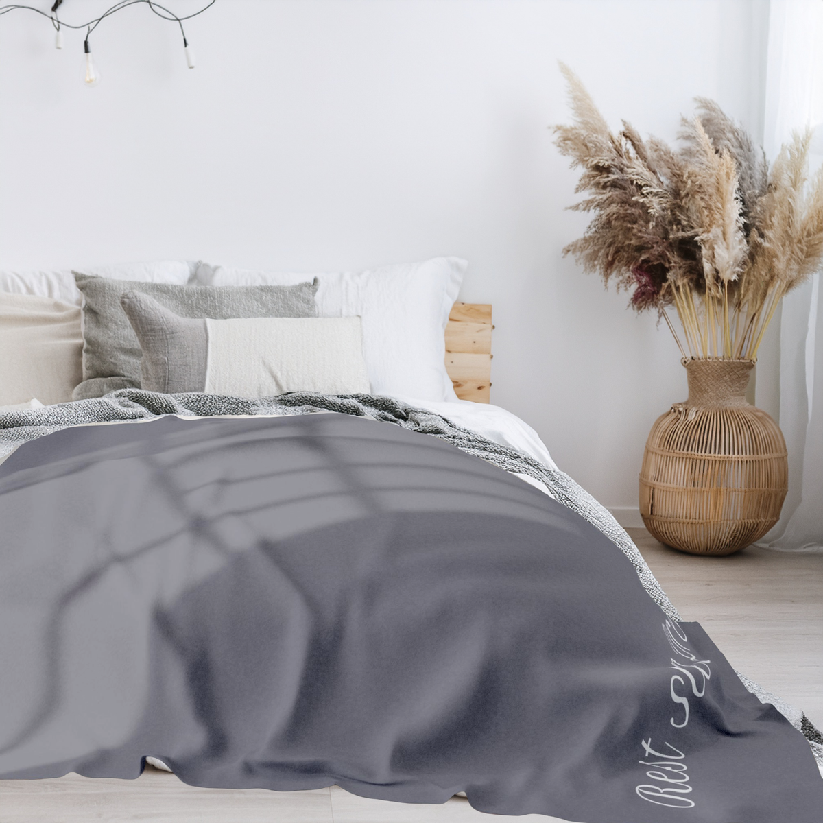 A gray throw blanket with white text 'Rest Sleep Dream' is draped over a bed in a bright, modern bedroom setting with a wooden headboard, neutral pillows, and a wicker vase with dried pampas grass.