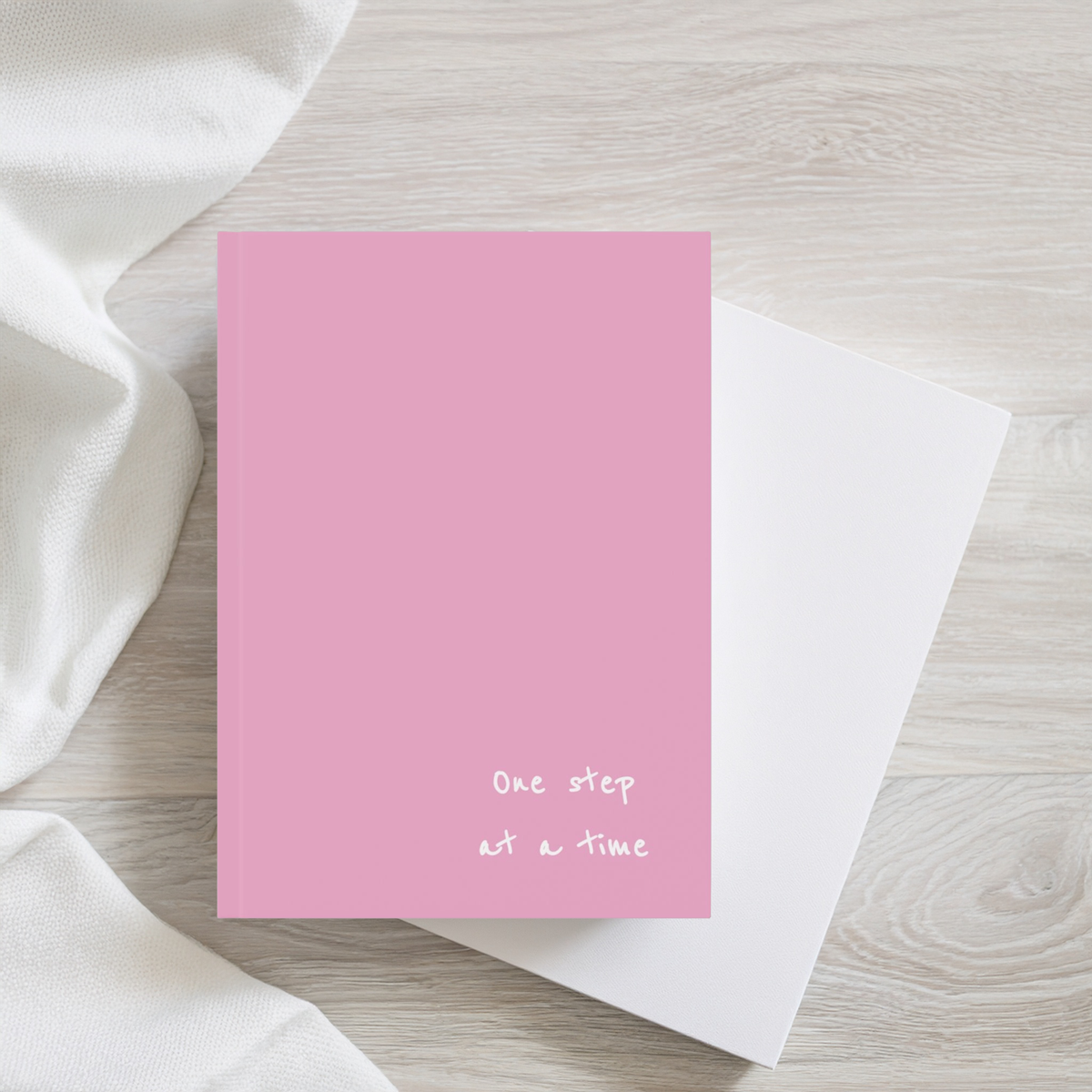 Pink a5 hardcover notebook journal with the phrase 'One step at a time' in small white script near the bottom, placed on top of a white notebook on a light wood surface with draped white fabric.