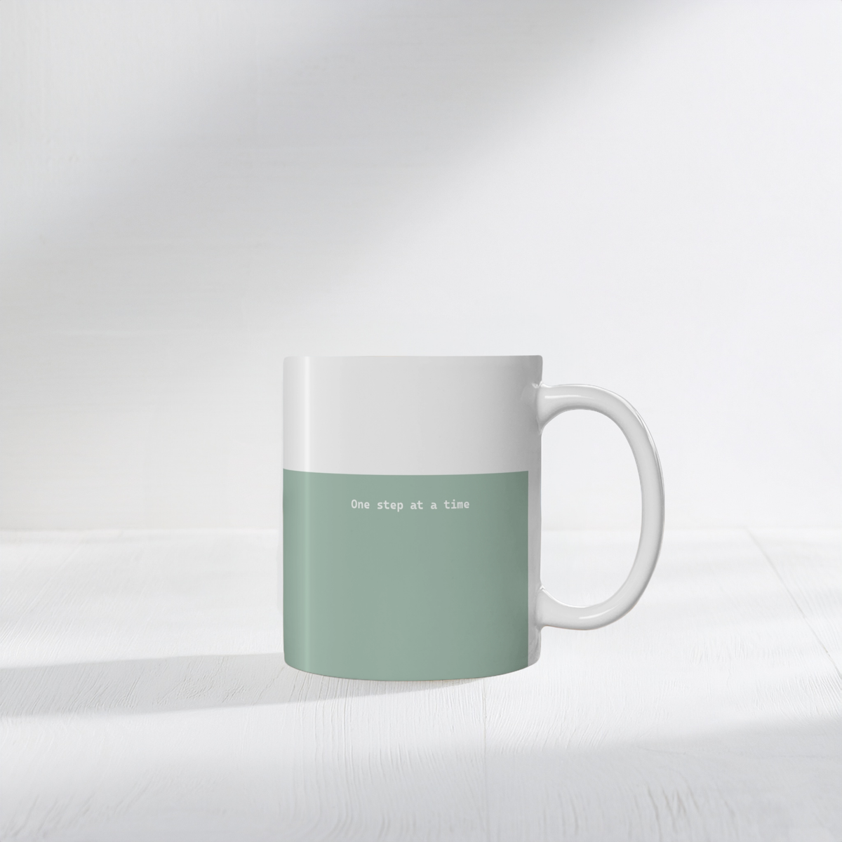 11oz green ceramic minimalist mug. Small white font in the middle ‘One step at a time’.  The mug sits on a white surface, minimalist background.