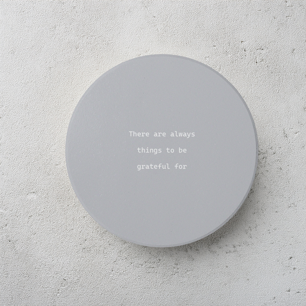 A round gray ceramic coaster with the printed phrase 'There are always things to be grateful for' in a white typewriter font, displayed on a textured light gray concrete surface.