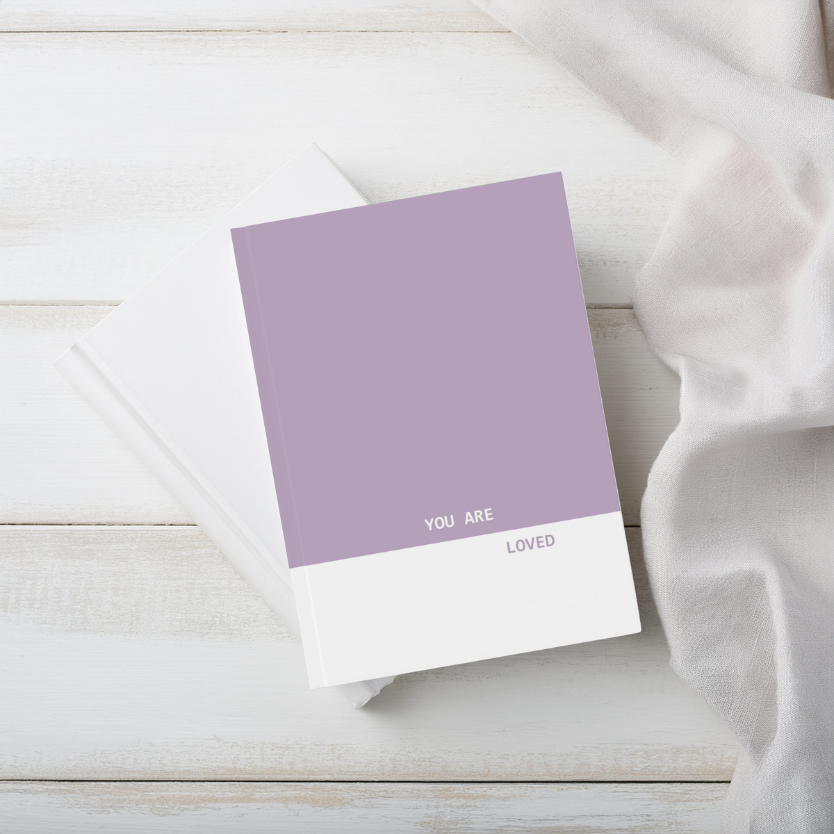 A purple and white a5 hardcover journal notebook, printed with the words 'YOU ARE LOVED' in white and purple, styled on a white wood surface with a white fabric napkin.