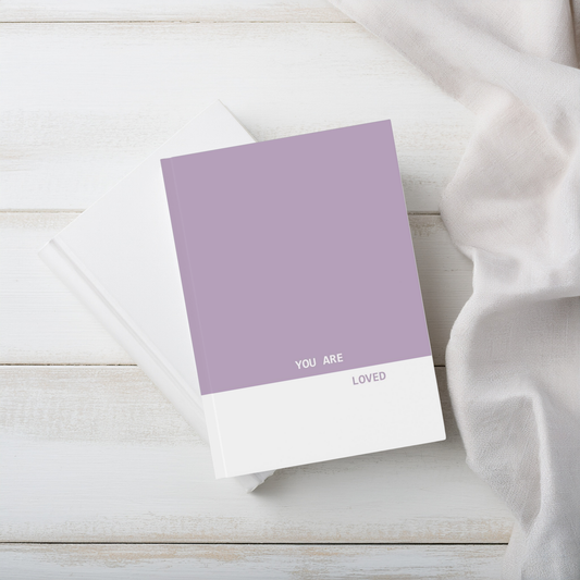 A purple and white a5 hardcover journal notebook, printed with the words 'YOU ARE LOVED' in white and purple, styled on a white wood surface with a white fabric napkin.