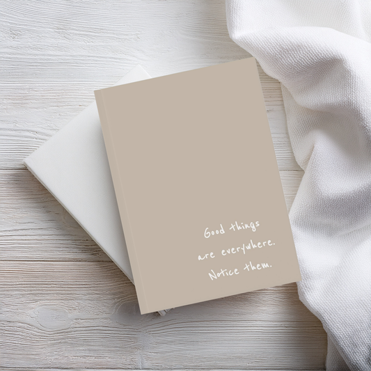 A beige brown a5 hardcover notebook with the handwritten quote 'Good things are everywhere. Notice them. The notebook sits on top of a plain white notebook on a wooden surface next to linen.
