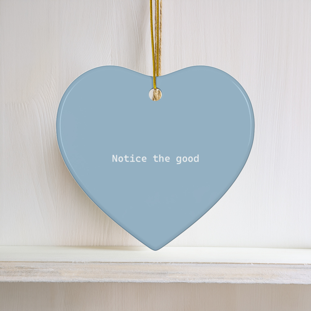 A light blue heart-shaped ceramic ornament hanging from a gold string, featuring the white printed text 'Notice the good' on a white wooden background.
