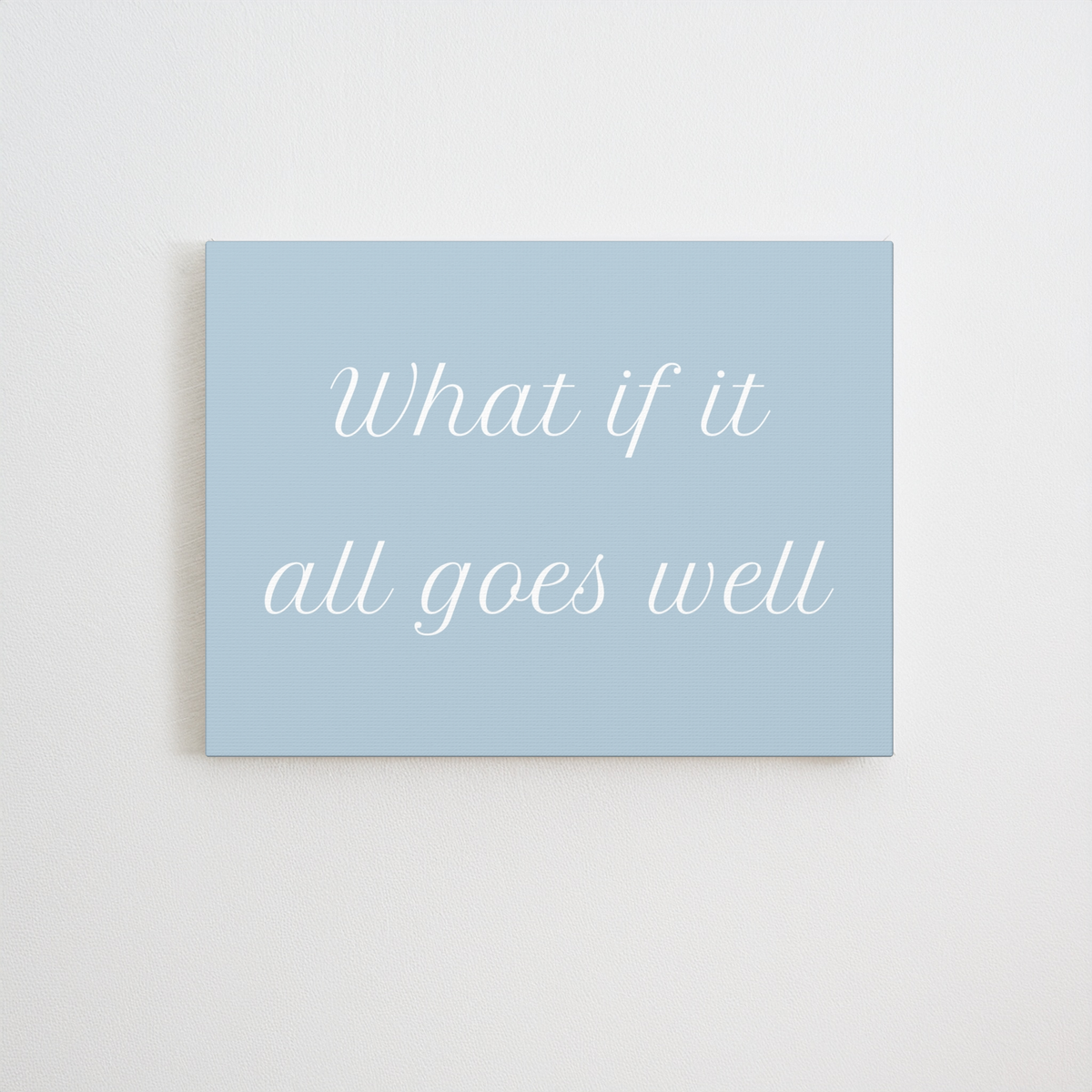 A canvas wall art print with the phrase 'What if it all goes well' in white cursive text on a light blue background, displayed against a neutral off-white wall.