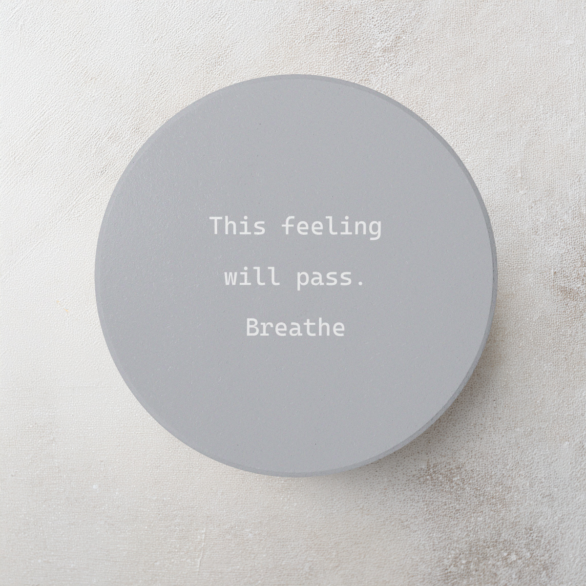 A round gray coaster with white typewriter-style text reading 'This feeling will pass. Breathe' on a light textured background.