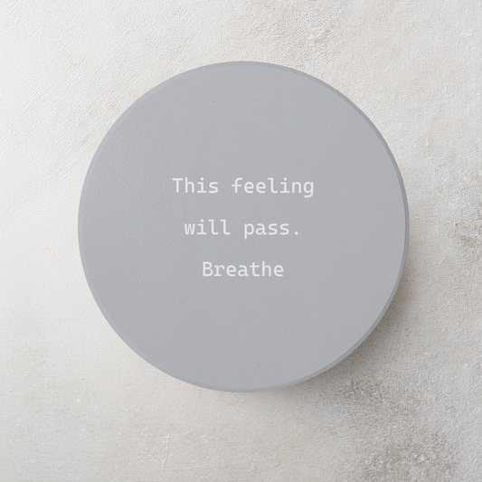 A round gray coaster with white typewriter-style text reading 'This feeling will pass. Breathe' on a light textured background.