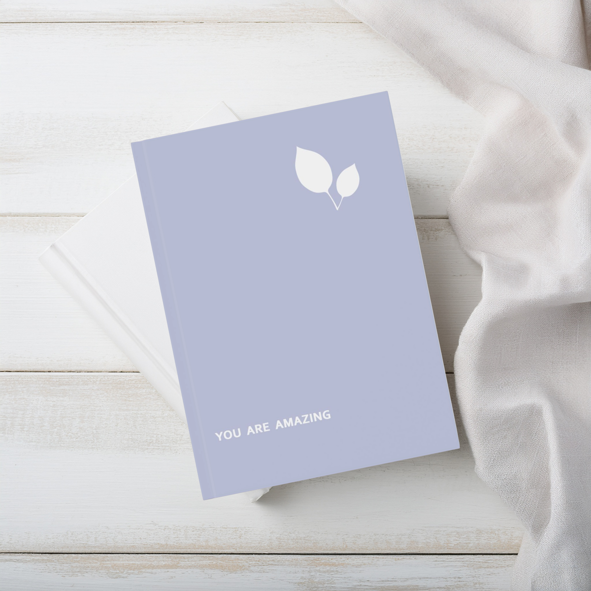 A lavender purple a5 hardcover notebook with a white leaf graphic and the words 'YOU ARE AMAZING' on the cover, styled on a white wooden surface next to a white fabric drape.