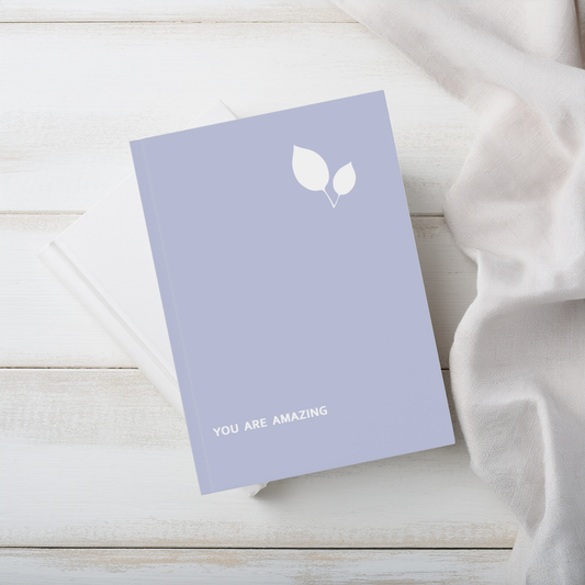 A lavender purple a5 hardcover notebook with a white leaf graphic and the words 'YOU ARE AMAZING' on the cover, styled on a white wooden surface next to a white fabric drape.