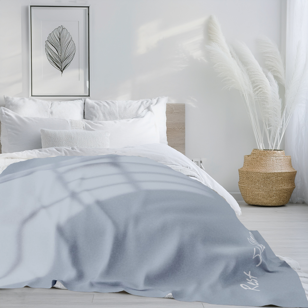 A light blue-gray throw blanket with white text, 'Rest Sleep Dream', is draped over a bed in a bright, modern bedroom. The scene includes white bedding, a wooden headboard, a framed leaf print, and a woven basket vase with pampas grass.