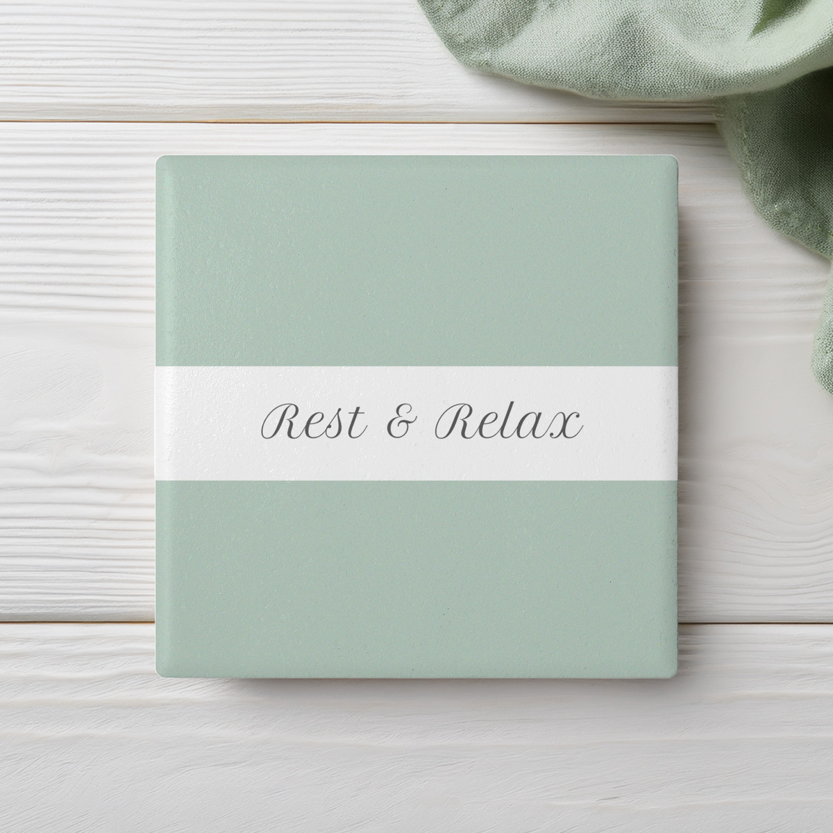 A square ceramic coaster with a pale sage green cover and a white horizontal band across the middle reading 'Rest & Relax' in gray script. It is photographed on a white wood surface with a soft green cloth partially visible in the top right corner.
