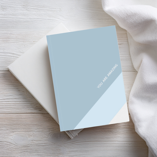 A5 hardcover journal notebook with blue large diagonal stripe and small white text 'You are amazing'. The notebook sits on top of a plain white notebook on a wooden surface next to a linen 