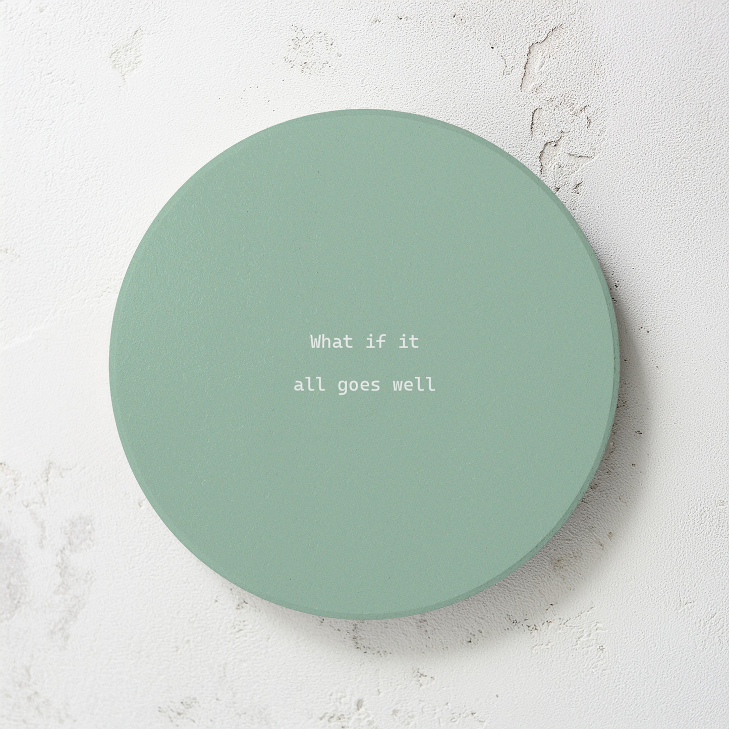 Green round ceramic coaster with modern style text ‘What if it all goes well’ in white font. The coaster is on a grey cement style surface.