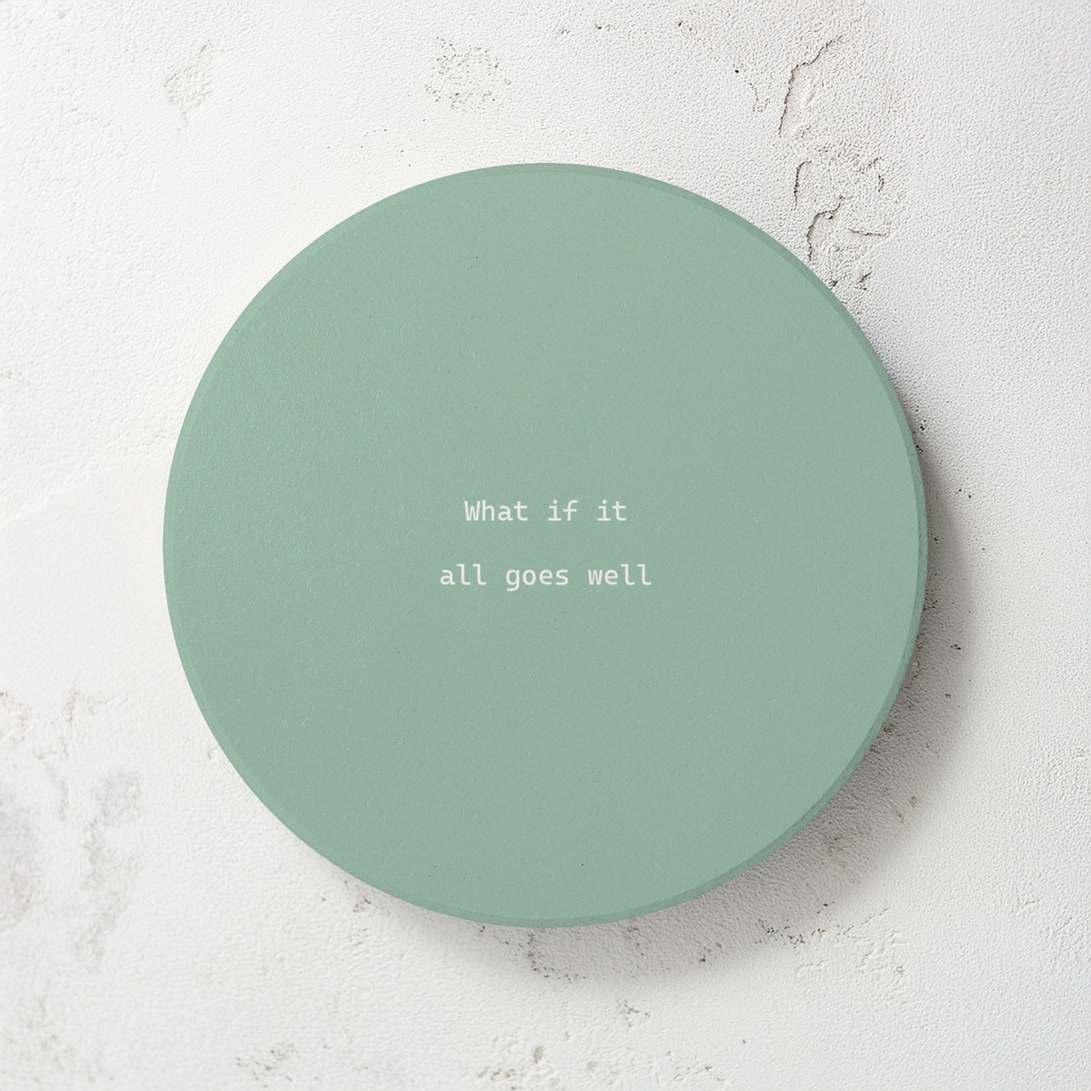 Green round ceramic coaster with modern style text ‘What if it all goes well’ in white font. The coaster is on a grey cement style surface.