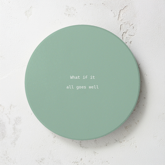 Green round ceramic coaster with modern style text ‘What if it all goes well’ in white font. The coaster is on a grey cement style surface.