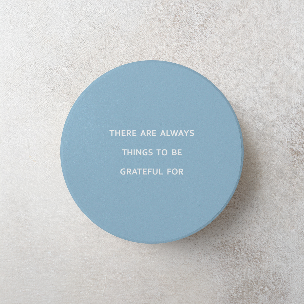 Blue round ceramic coaster with modern style text ‘There are always things to be grateful for’ in white font. The coaster is on a grey cement style surface.