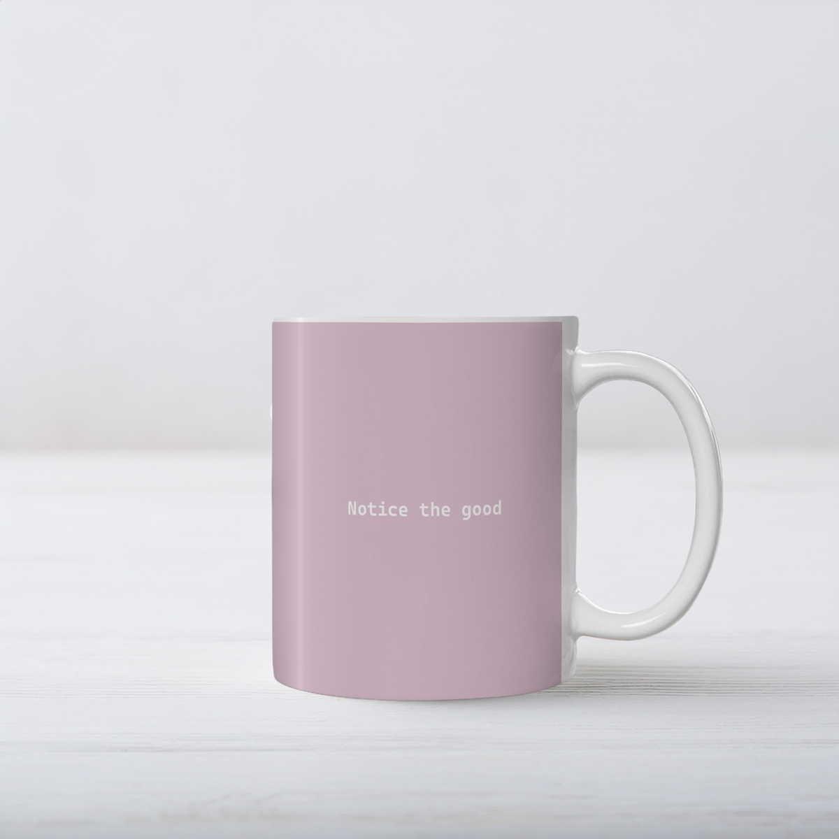 Pale pink ceramic mug with small white font in the middle ‘Notice the good'. The mug sits on a white wooden surface, minimalist background.