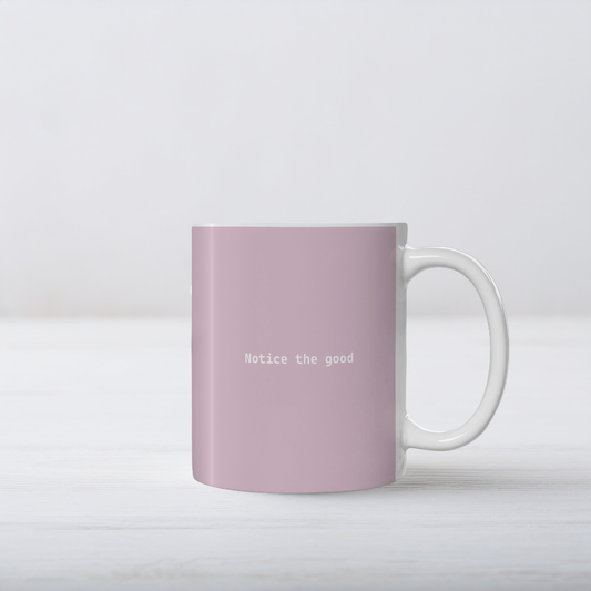 Pale pink ceramic mug with small white font in the middle ‘Notice the good'. The mug sits on a white wooden surface, minimalist background.