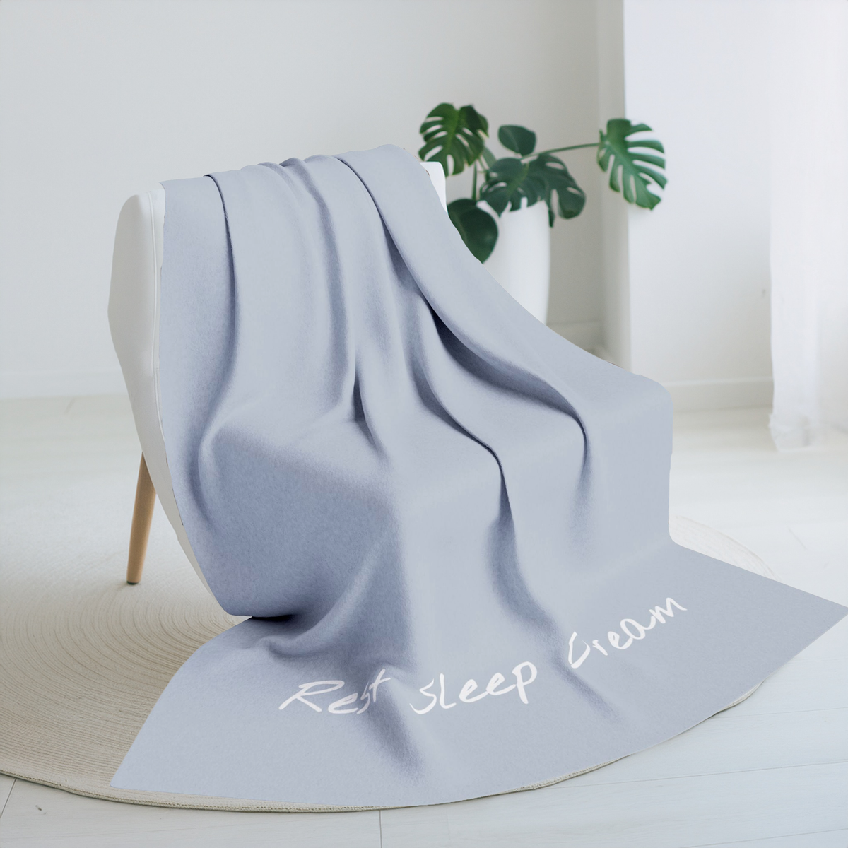 Pale blue blanket with large font in white for the text ‘Rest Sleep Dream’ along the bottom.  The blanket is on a chair in a living room with a plant in the background.