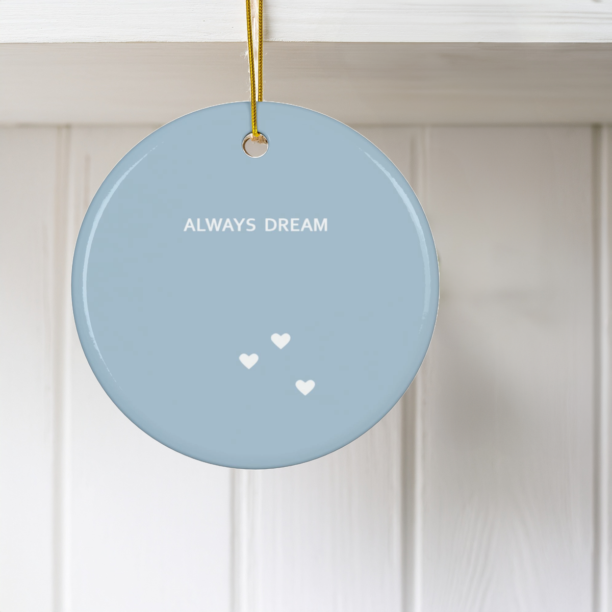 A round light blue ceramic Christmas ornament hanging from a gold string against a white paneled wall. The ornament features the text 'ALWAYS DREAM' in white, with three small white hearts below.
