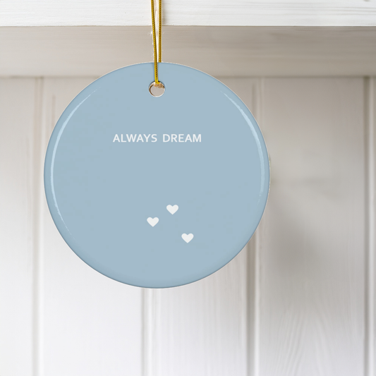 A round light blue ceramic Christmas ornament hanging from a gold string against a white paneled wall. The ornament features the text 'ALWAYS DREAM' in white, with three small white hearts below.