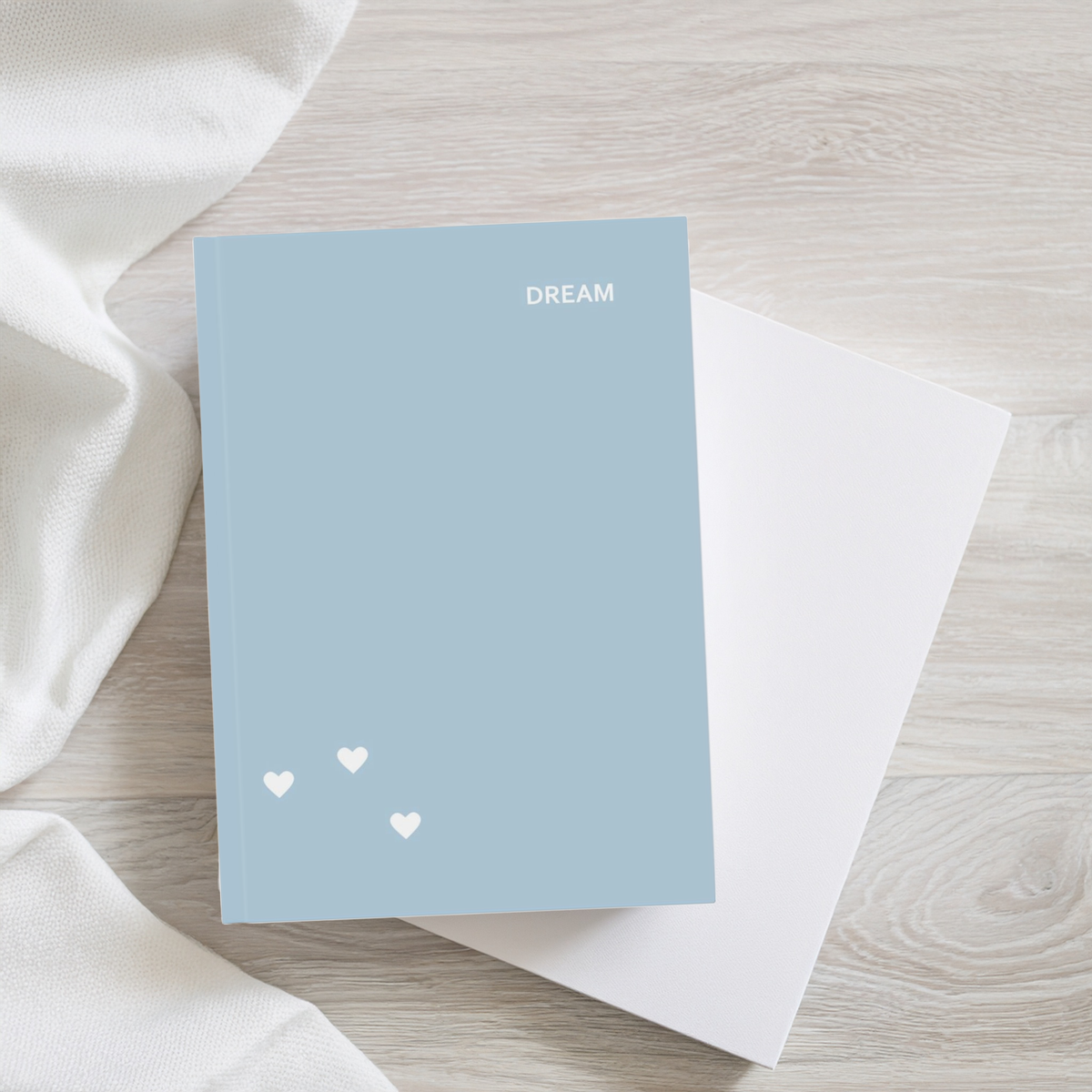 A light blue hardcover notebook with the word 'DREAM' in white at the top right and three small white hearts near the bottom left, styled on a light wood surface with a white fabric prop and a white notebook underneath.