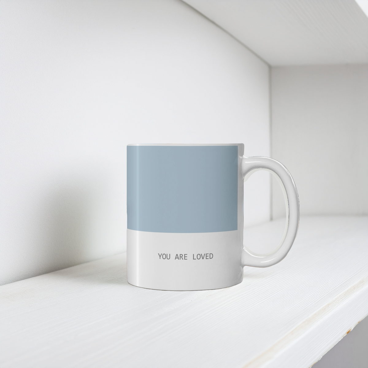 11oz blue and white ceramic minimalist mug. Small font at the bottom ‘you are loved’.  The mug sits on a white wooden shelf, minimalist background.