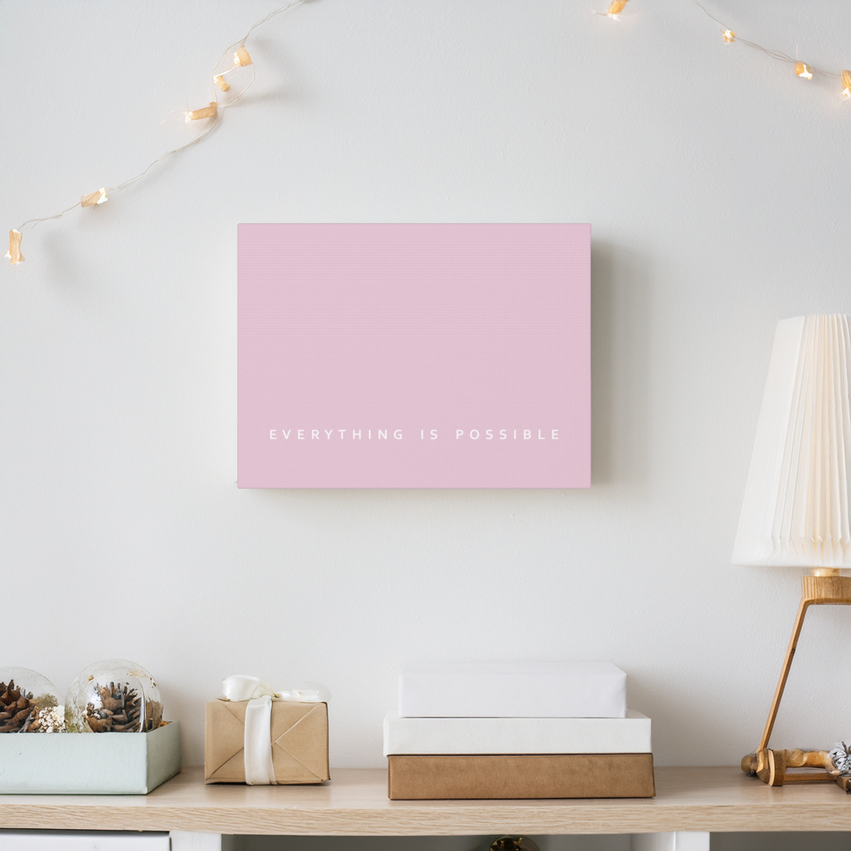 A minimalist pink canvas wall art print with the white text 'EVERYTHING IS POSSIBLE' displayed on a light gray wall above a styled wooden console table with a lamp, gift boxes, and decorative items.