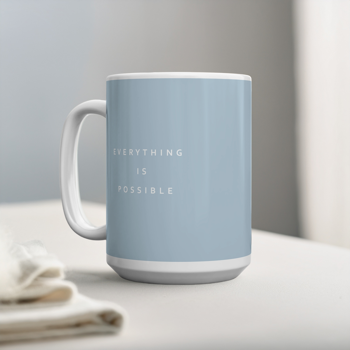 Blue ceramic mug with small white font in the middle ‘Everything is possible’. The mug sits on a white table, next to some linen, minimalist background.