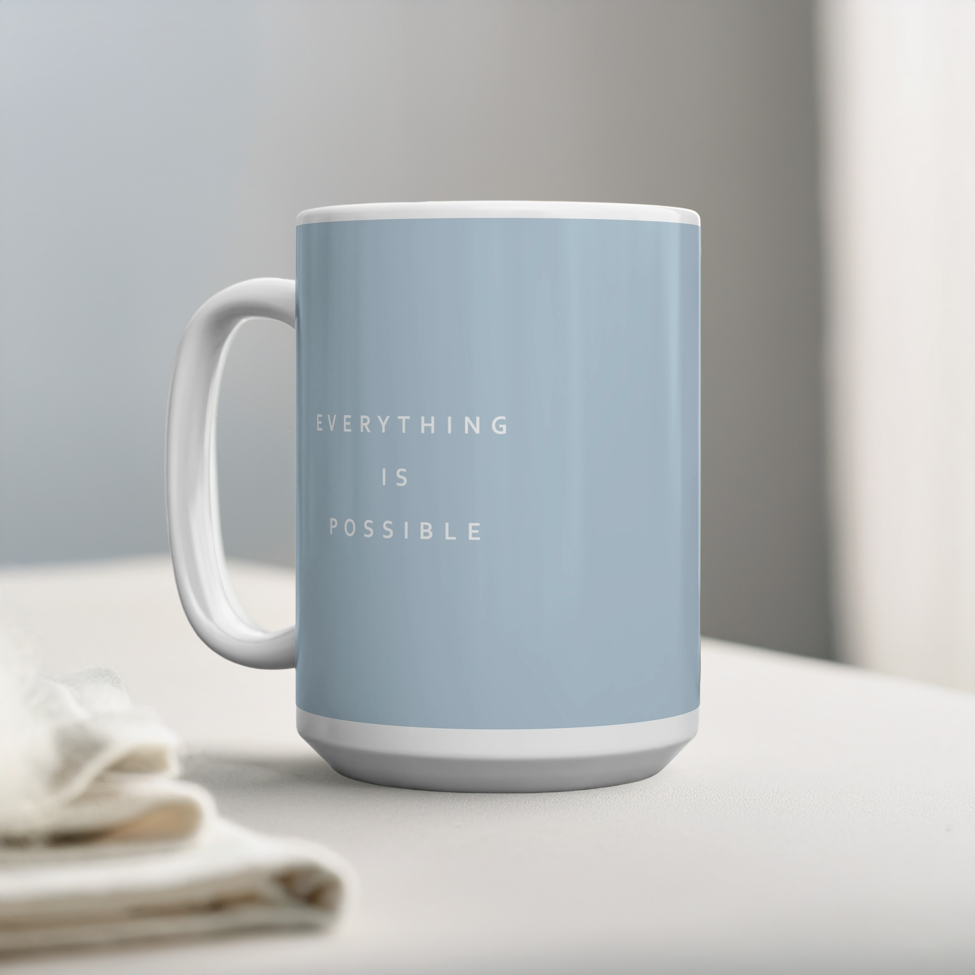 Blue ceramic mug with small white font in the middle ‘Everything is possible’. The mug sits on a white table, next to some linen, minimalist background.