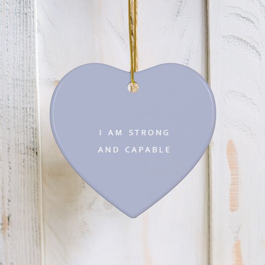 A heart-shaped ceramic ornament in a dusty lavender-blue color, hanging from a gold string against a white wooden door. The ornament features the text 'I AM STRONG AND CAPABLE' in white capital letters.