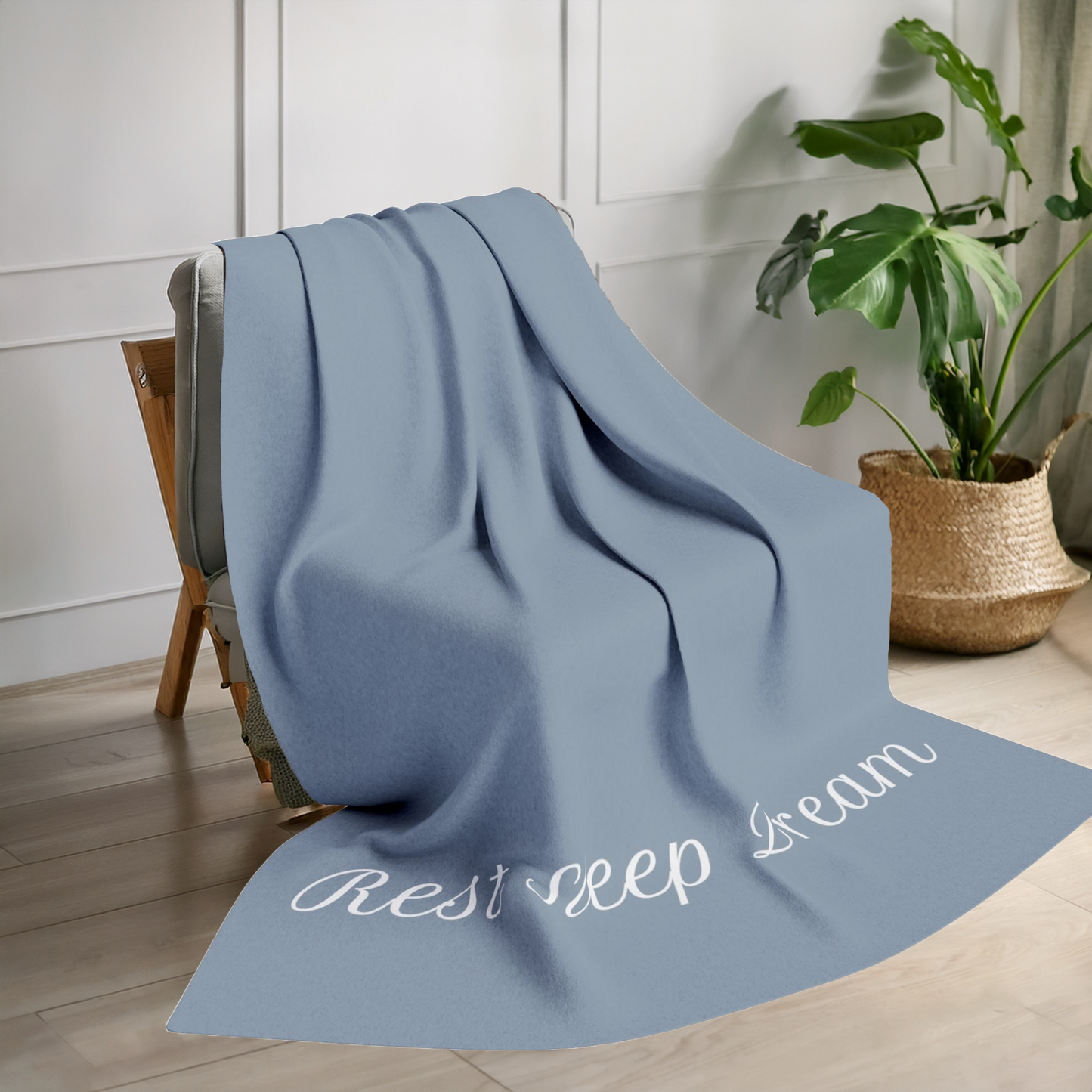 Blue blanket with large, elegant font in white for the text ‘Rest Sleep Dream’ along the bottom.  The blanket is on a chair in a living room with a plant in the background.